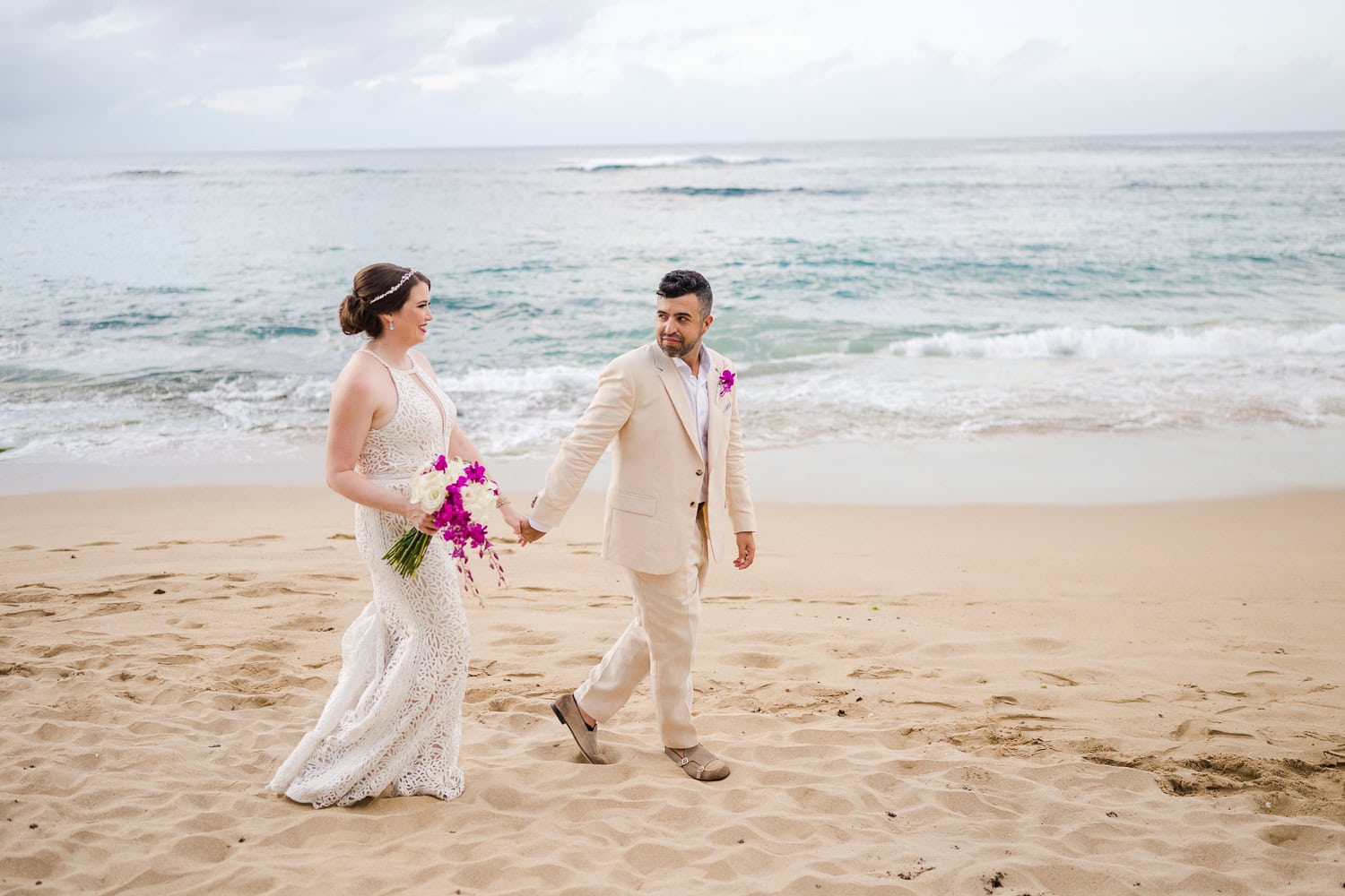 Bride and groom walking hand in hand along the sandy beach at Villa Montana Beach Resort in Puerto Rico, with the ocean waves gently crashing in the background.