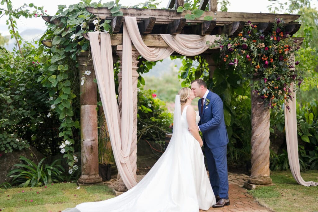 Bride and groom sharing a kiss under a floral-draped pergola at Hacienda Siesta Alegre, surrounded by lush greenery and vibrant floral arrangements in Puerto Rico