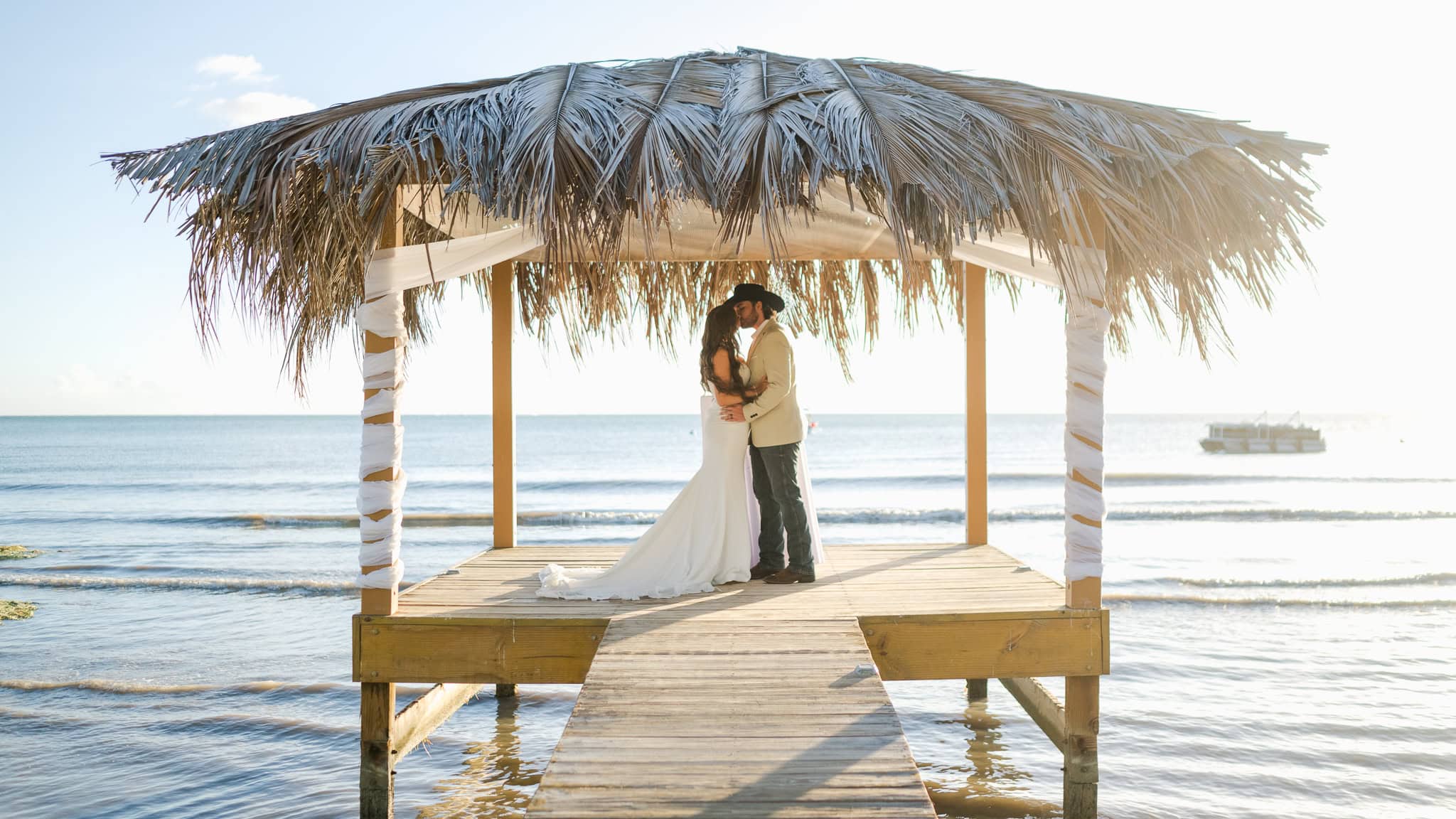 Romantic Caribbean beach wedding photo of a bride and groom kissing under a thatched-roof gazebo at sunset