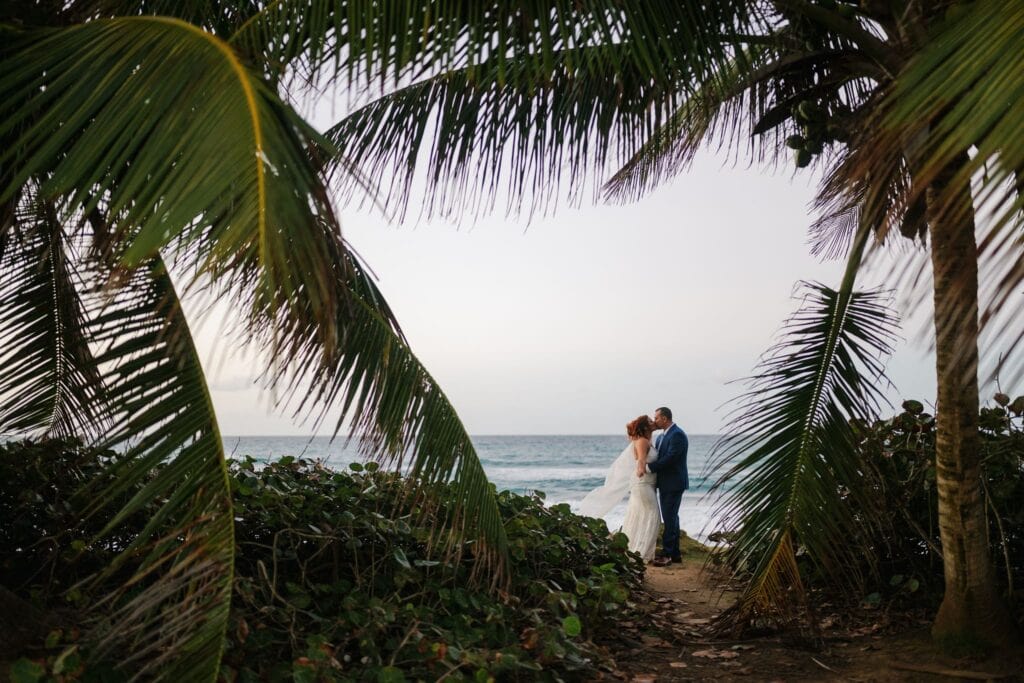 Bride and groom standing under a palm tree at Escambrón Beach, Caribbean Sea visible in the background at sunset