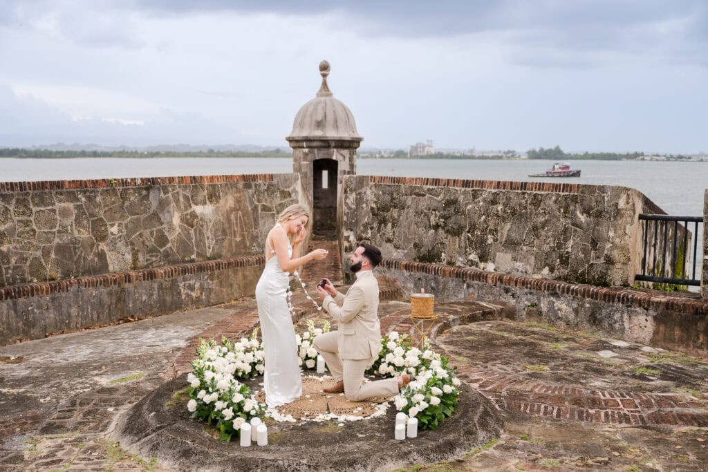 Groom on one knee proposing to bride on the Bastión de San Agustín with the San Juan bay behind them