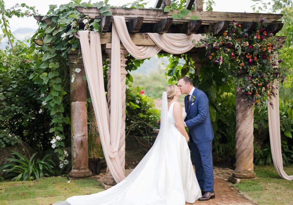 Bride and groom sharing a kiss under a floral-draped pergola at Hacienda Siesta Alegre, surrounded by lush greenery and vibrant floral arrangements in Puerto Rico